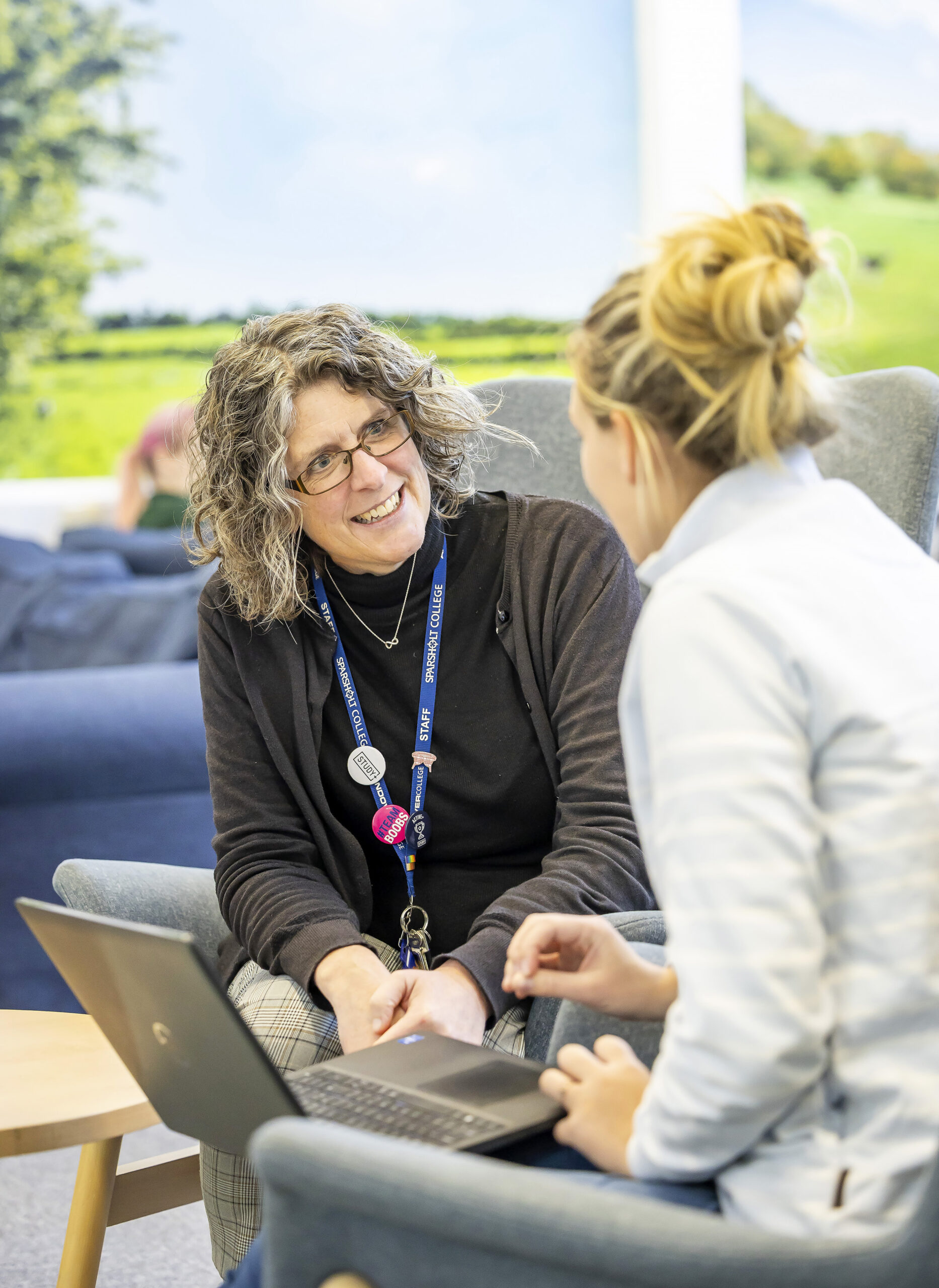 Two people seated in armchairs having a discussion while looking at a laptop. One person is wearing a lanyard with multiple badges, and the setting is a bright, casual indoor space with a scenic countryside mural in the background